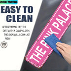 Hand wiping pink vintage tin sign reading 'THE PINK PALACE' with blue cloth, easy to clean.